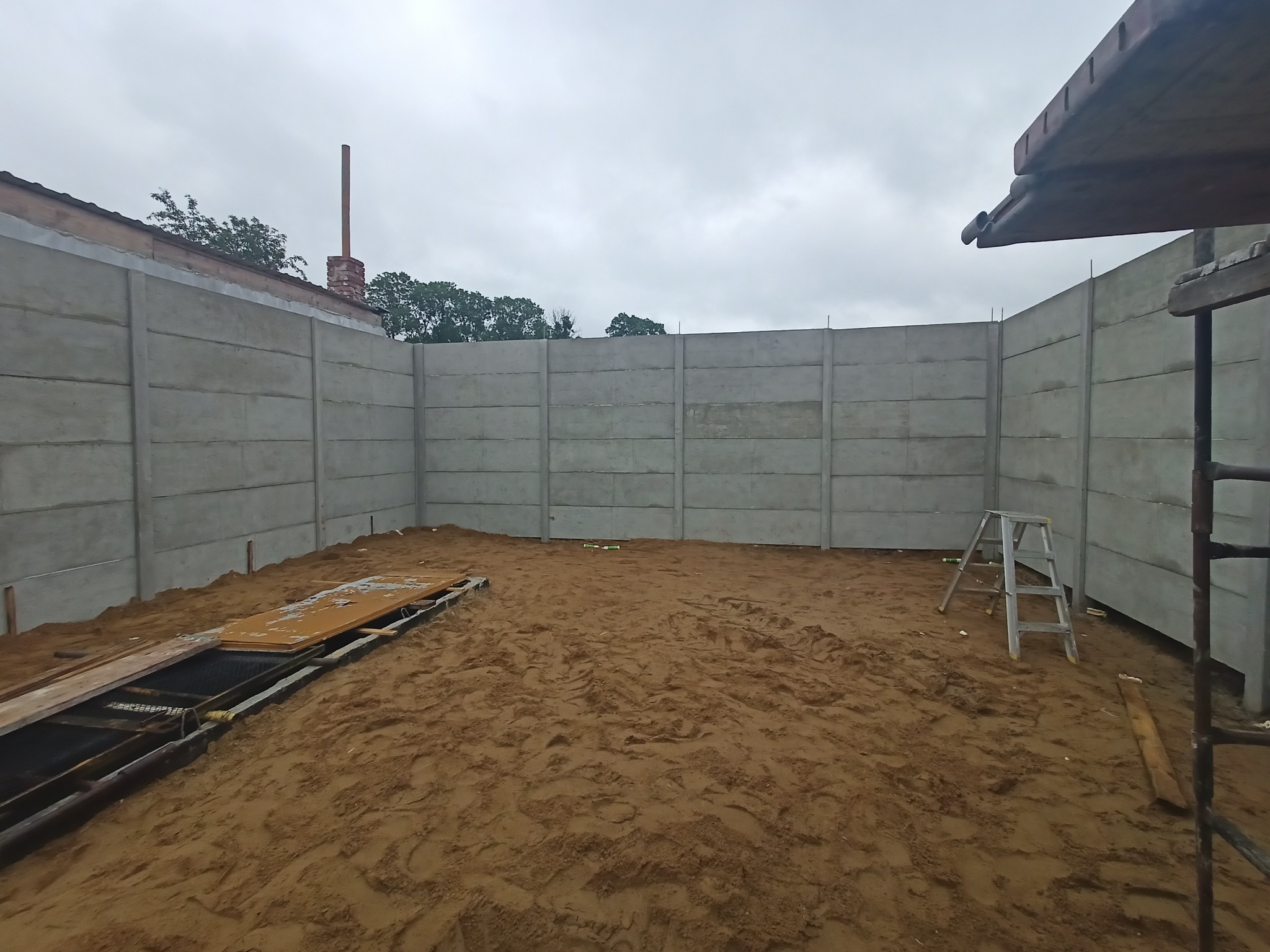 Wysoka, prefabrykowana betonowa ściana ogrodzeniowa w trakcie montażu, widoczne elementy konstrukcyjne i podłoże z piasku, drabina oparta o ścianę.