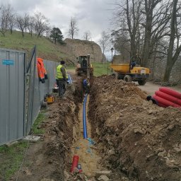 Greenking - Wykop z ułożoną niebieską rurą, robotnicy w odblaskowych kamizelkach, koparka i wywrotka w tle, zabezpieczony teren budowy, widok na wzgórze z ruinami zamku.