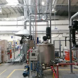 Instalacja przemysłowa z rurociągami, zbiornikiem ze stali nierdzewnej i panelem sterowania w hali produkcyjnej z żółtymi liniami na podłodze.