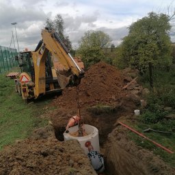 Greenking - Żółta koparko-ładowarka CAT umieszcza betonowy krąg w wykopie, dwóch pracowników w trakcie instalacji przyłącza, widoczna rura kanalizacyjna w ziemi.