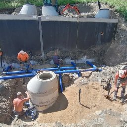 Greenking - Montaż dwukomorowego betonowego zbiornika na szambo z niebieskimi rurami kanalizacyjnymi i pracującymi robotnikami w pomarańczowych koszulkach, widoczny mini-koparka na górze wykopu.