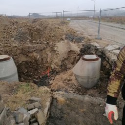 Greenking - Montaż betonowych studzienek kanalizacyjnych w wykopie, widoczny fragment ogrodzenia placu budowy i odległy krajobraz w pochmurny dzień.