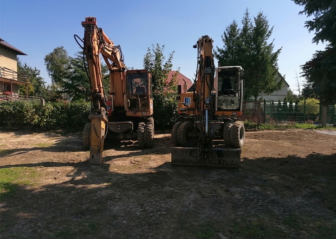 Dwie pomarańczowe koparki na placu budowy, jedna z młotem hydraulicznym, druga z łyżką, widoczne budynki w tle.