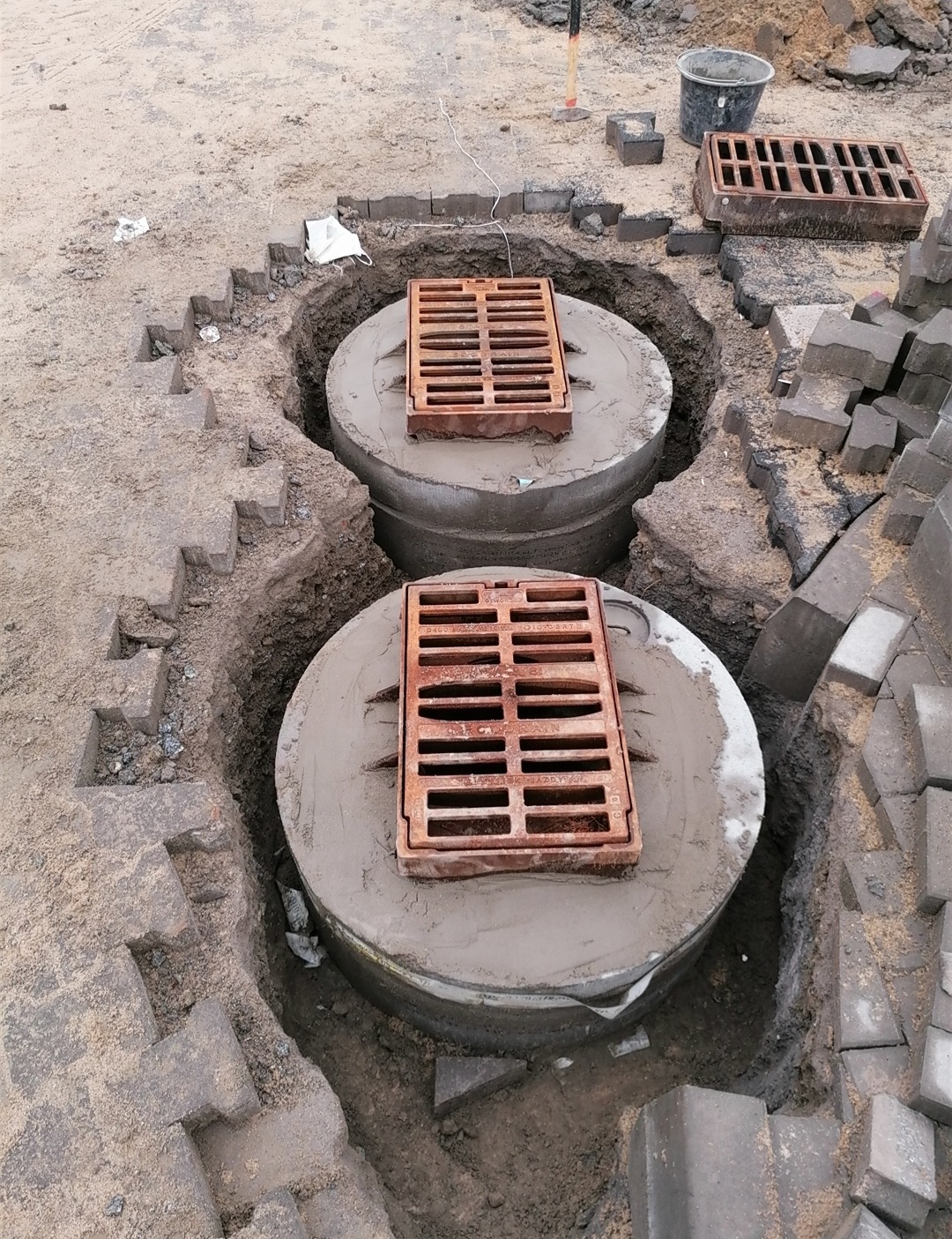 Dwa betonowe kręgi studni kanalizacyjnej z żeliwnymi kratami w wykopie, częściowo obsypane ziemią i otoczone kostką brukową, widoczny fragment maski ochronnej.