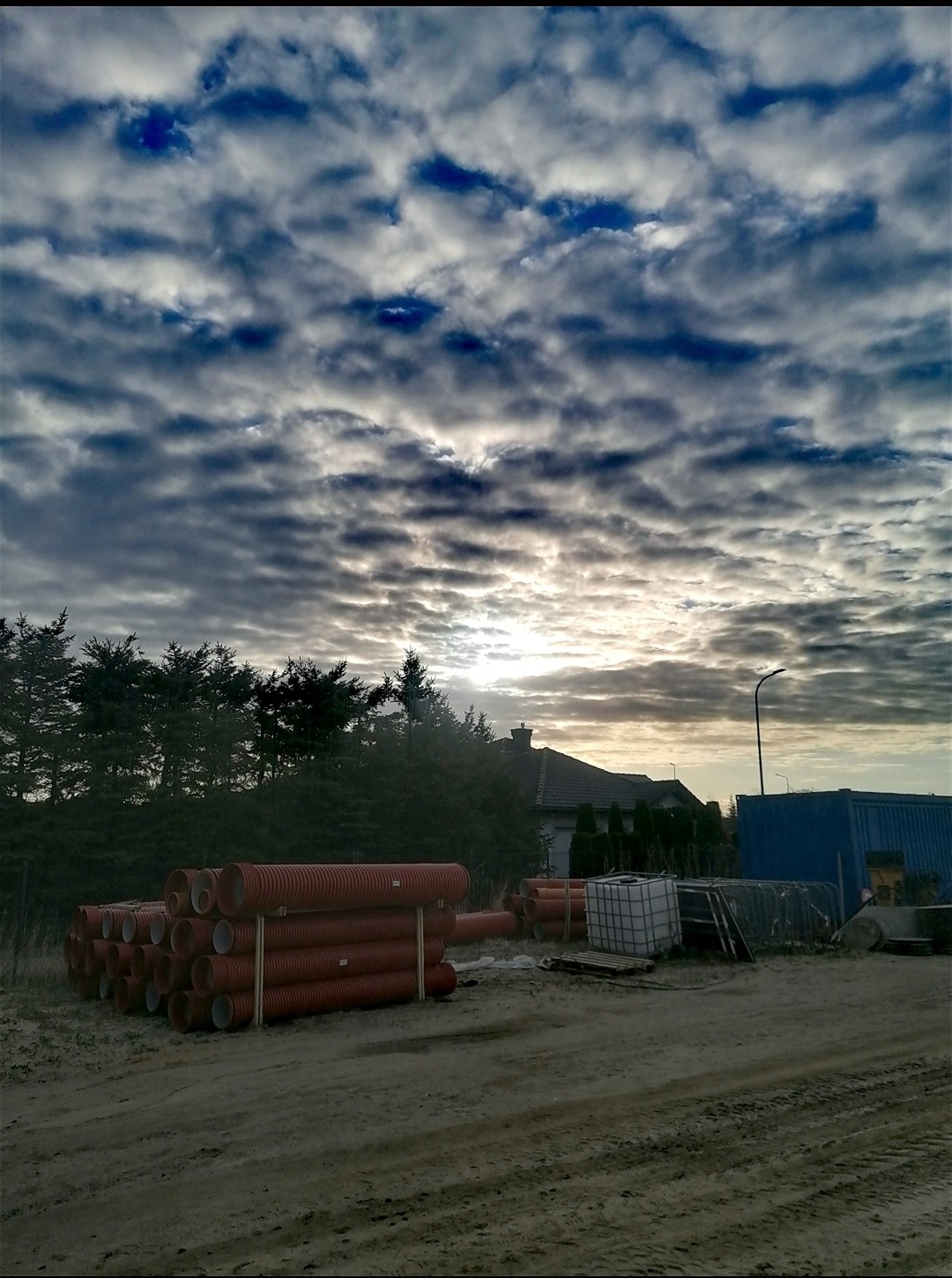 Stos rur drenarskich z tworzywa sztucznego na placu budowy, w tle drzewa, dom i zachmurzone niebo.