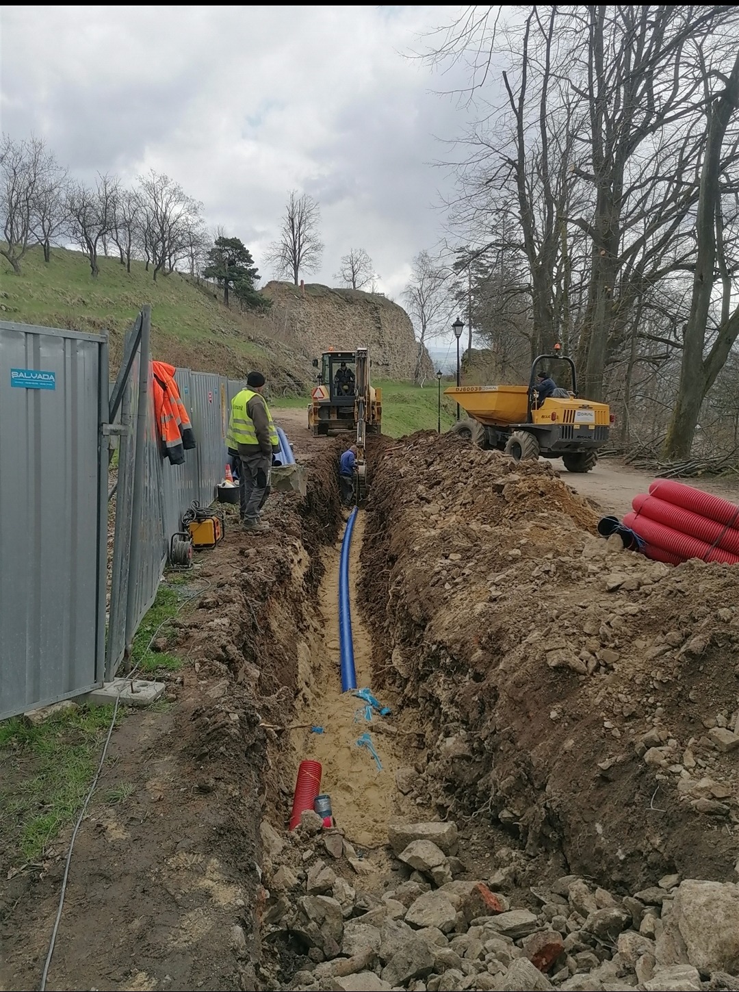 Wykop z ułożoną niebieską rurą, robotnicy w odblaskowych kamizelkach, koparka i wywrotka w tle, zabezpieczony teren budowy, widok na wzgórze z ruinami zamku.