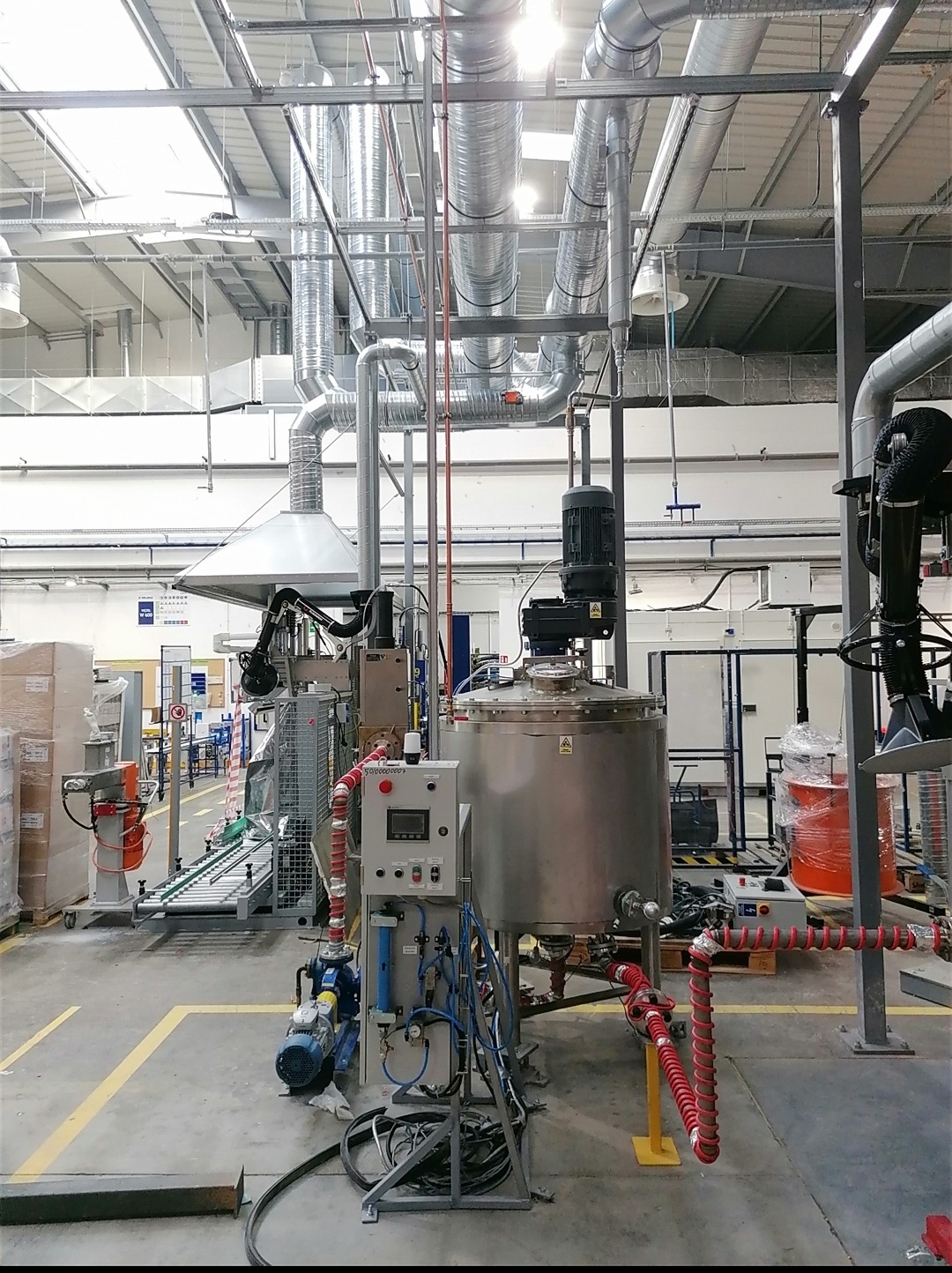 Instalacja przemysłowa z rurociągami, zbiornikiem ze stali nierdzewnej i panelem sterowania w hali produkcyjnej z żółtymi liniami na podłodze.