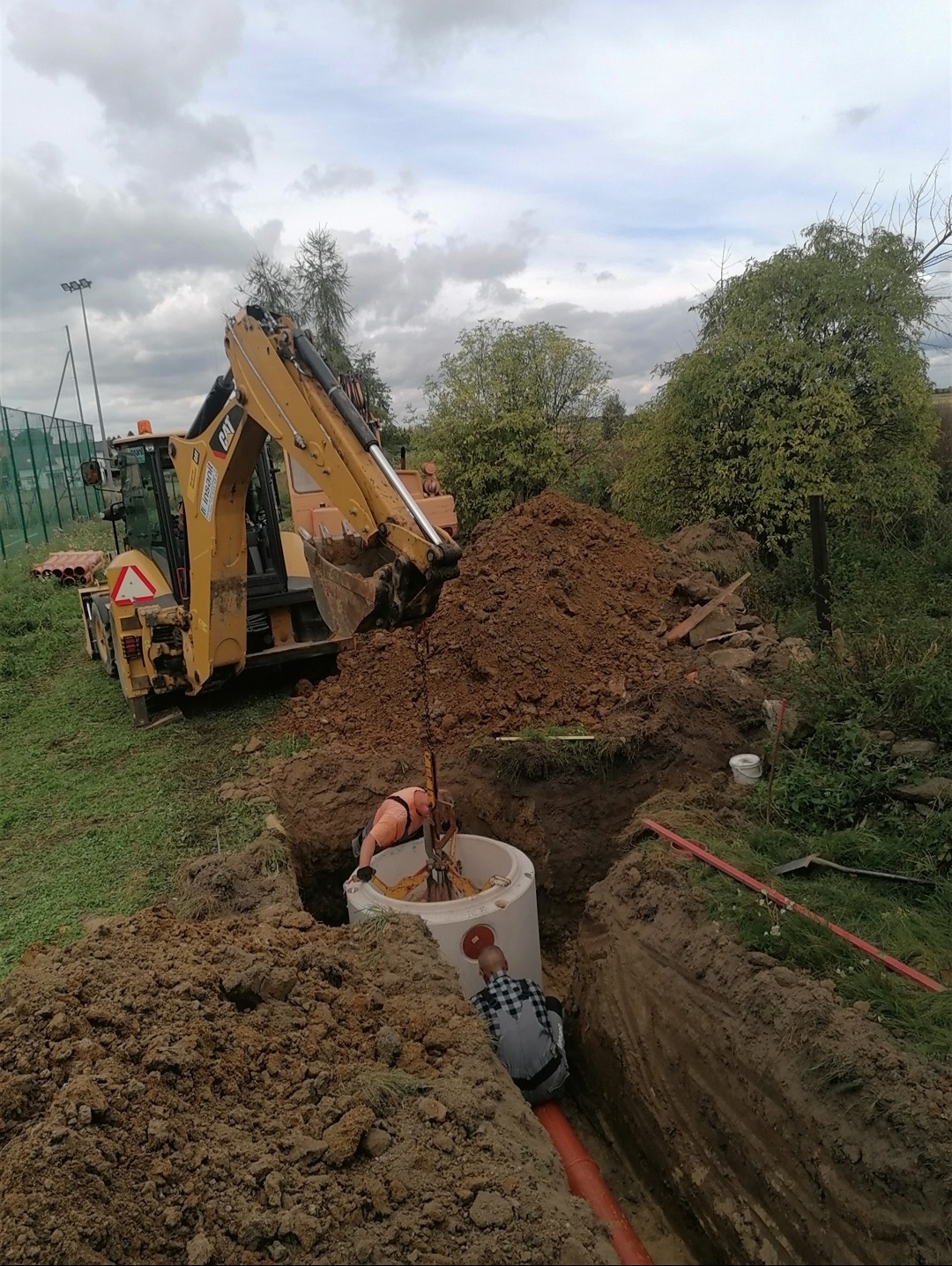 Żółta koparko-ładowarka CAT umieszcza betonowy krąg w wykopie, dwóch pracowników w trakcie instalacji przyłącza, widoczna rura kanalizacyjna w ziemi.