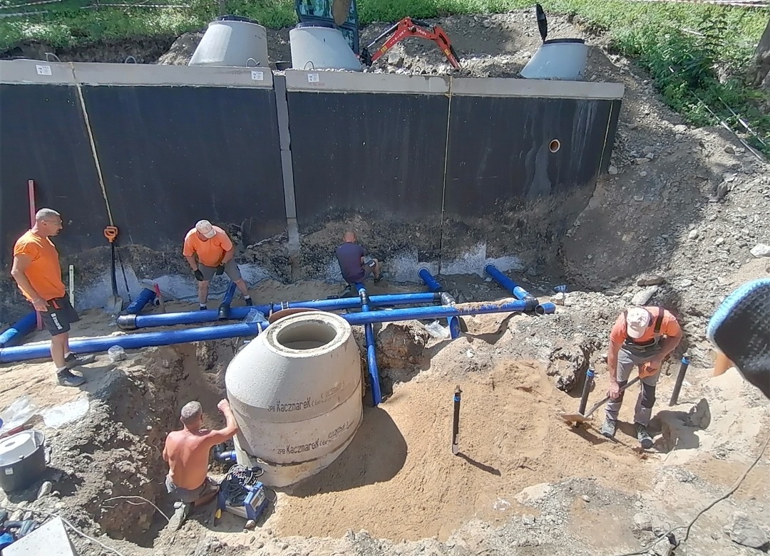 Montaż dwukomorowego betonowego zbiornika na szambo z niebieskimi rurami kanalizacyjnymi i pracującymi robotnikami w pomarańczowych koszulkach, widoczny mini-koparka na górze wykopu.