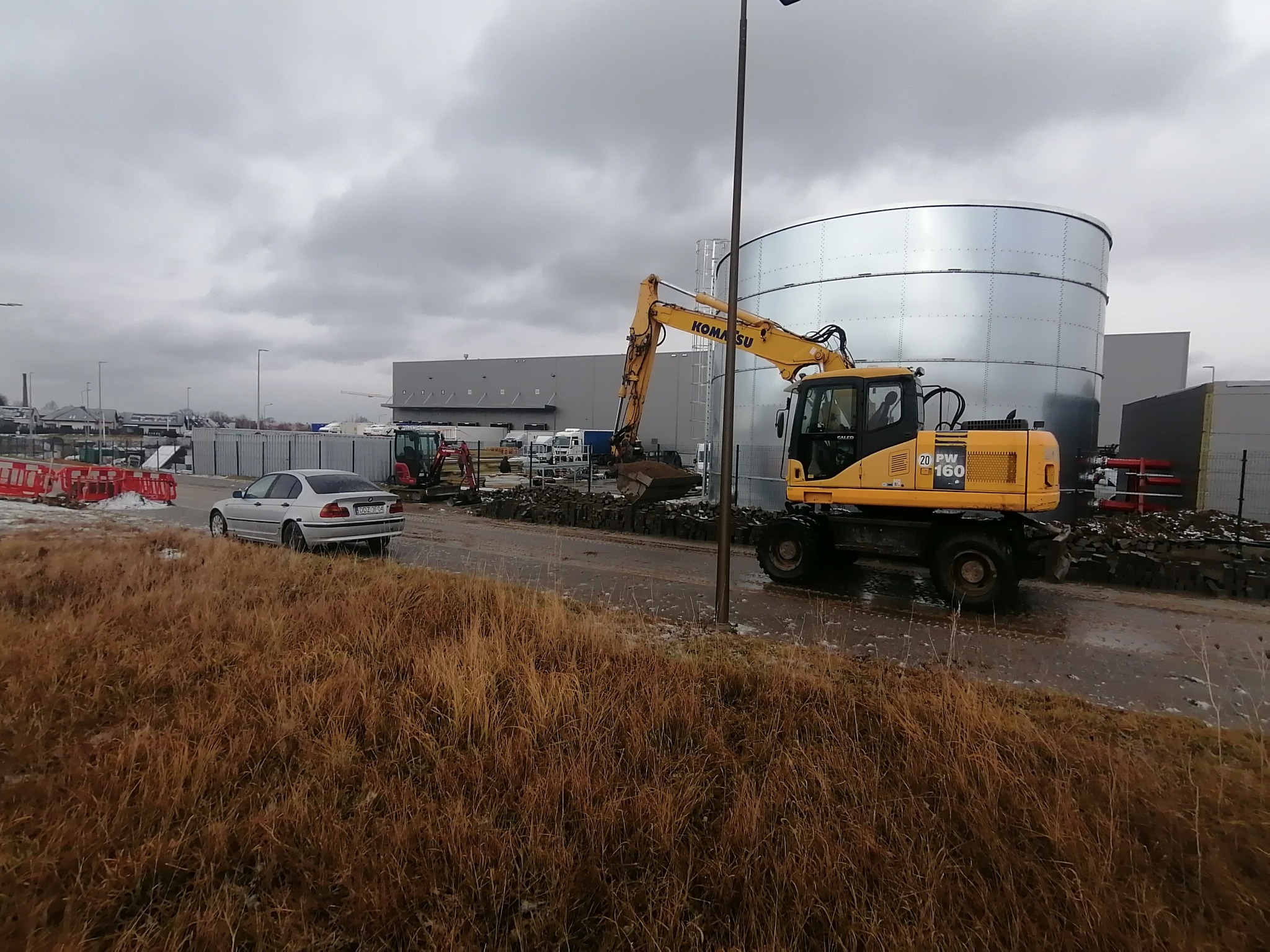 Żółta koparka Komatsu PW160 na placu budowy, widoczny srebrny zbiornik, w tle budynek magazynowy i samochód osobowy, pochmurne niebo.