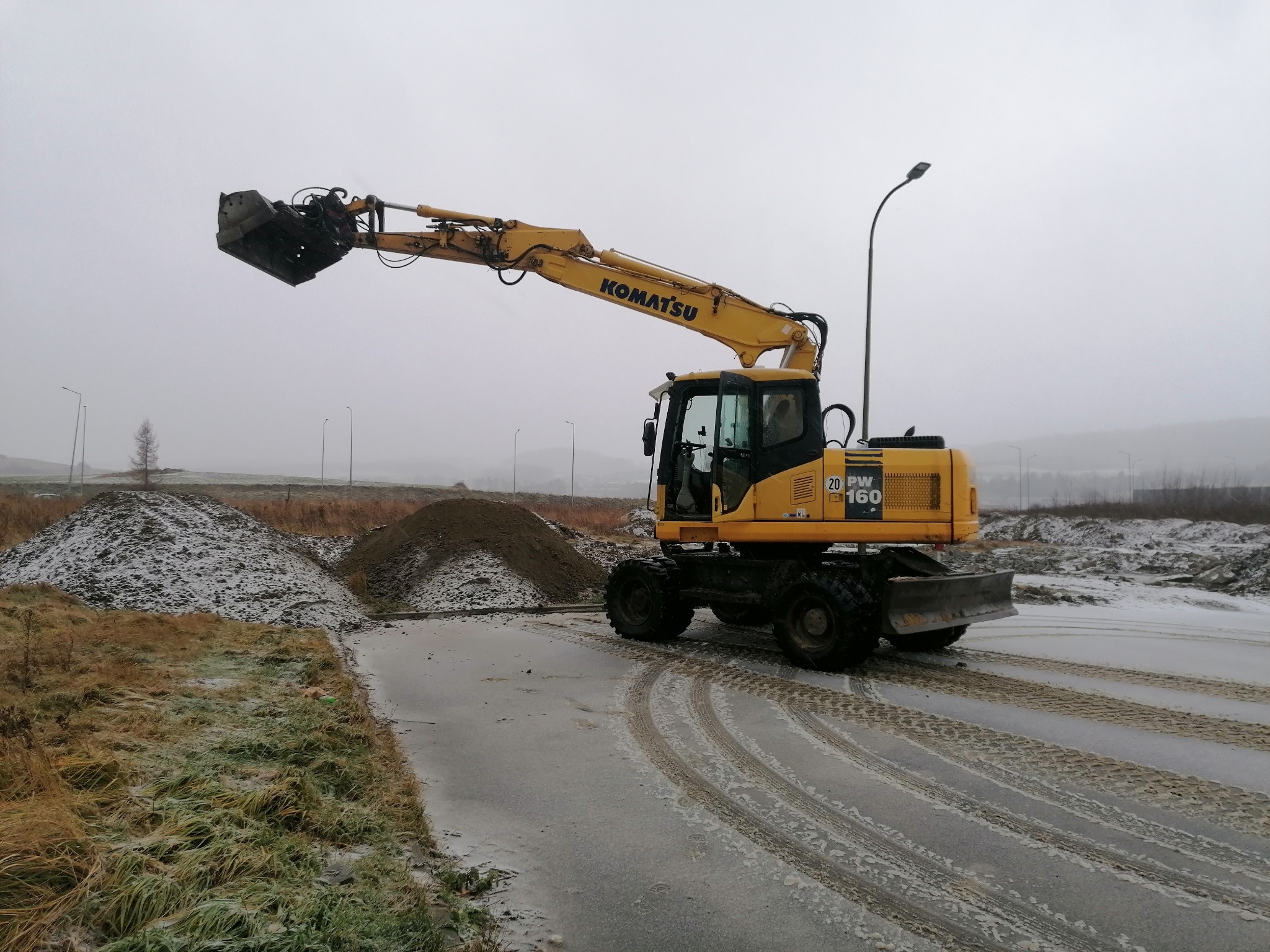 Żółta koparka kołowa Komatsu PW160 na placu budowy w pochmurny dzień, z widocznymi śladami opon na mokrej nawierzchni i hałdami ziemi w tle.