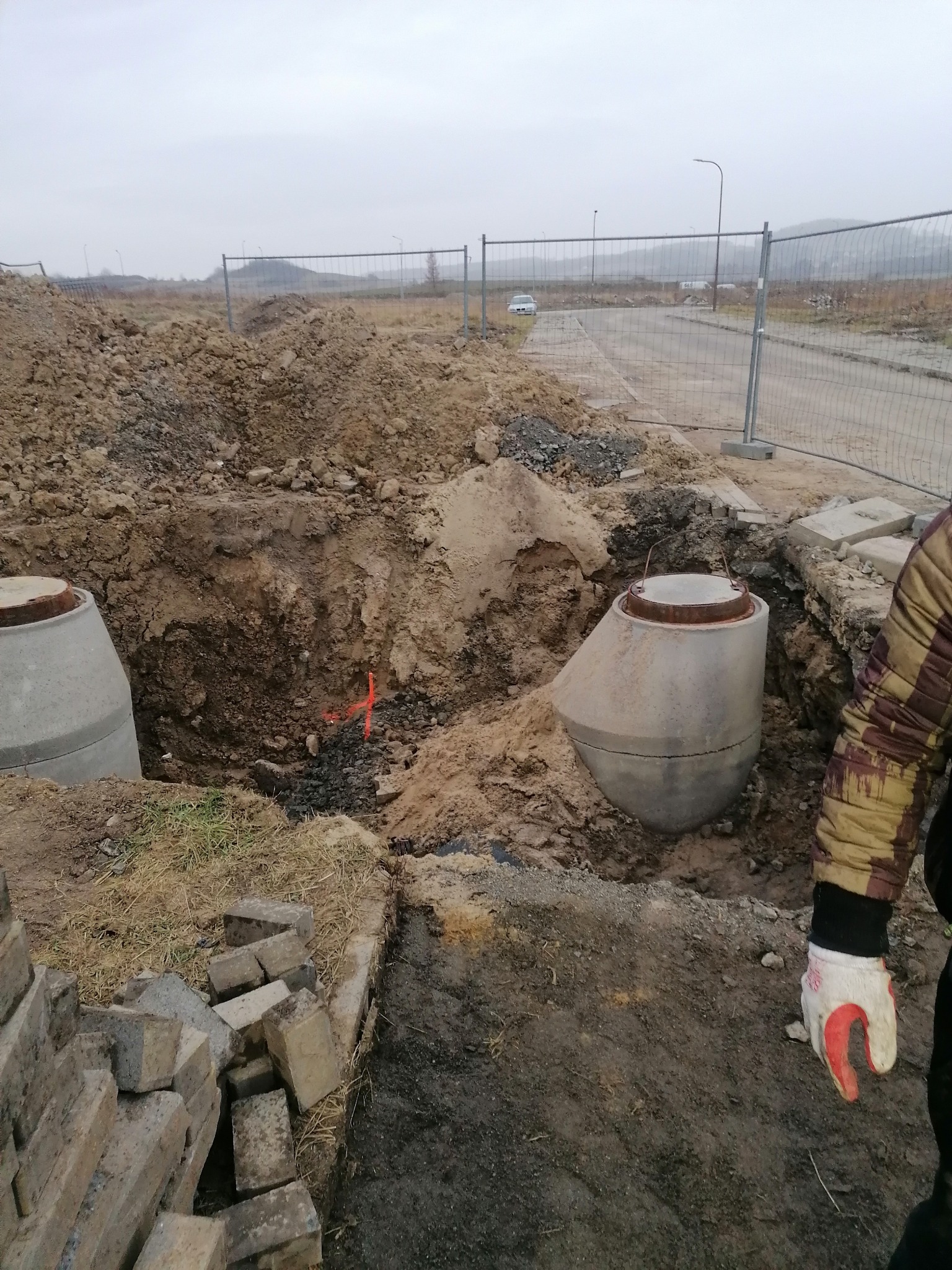 Montaż betonowych studzienek kanalizacyjnych w wykopie, widoczny fragment ogrodzenia placu budowy i odległy krajobraz w pochmurny dzień.