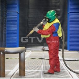 SAP Sp. z o.o. - Pracownik w kombinezonie ochronnym i hełmie śrutuje metalowy element w komorze śrutowniczej z niebieskimi panelami.