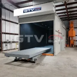 Wnętrze hali z komorą śrutowniczą GTV Blast z otwartą bramą i wysuwanym stołem roboczym, widoczna instalacja wentylacyjna i filtracja powietrza.