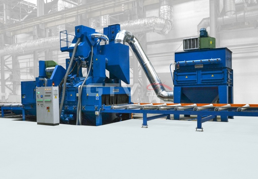 Niebieska, przemysłowa oczyszczarka rolkowa z transportem rolkowym, widoczny panel sterowania i rura odprowadzająca, na tle hali produkcyjnej.