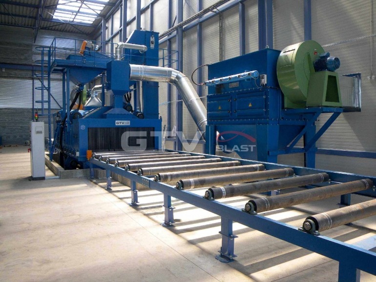 Kompleksowa linia technologiczna do obróbki strumieniowo-ściernej w hali produkcyjnej, z elementami podawczymi w postaci rolek transportowych i systemem odpylania z zielonym wentylatorem.