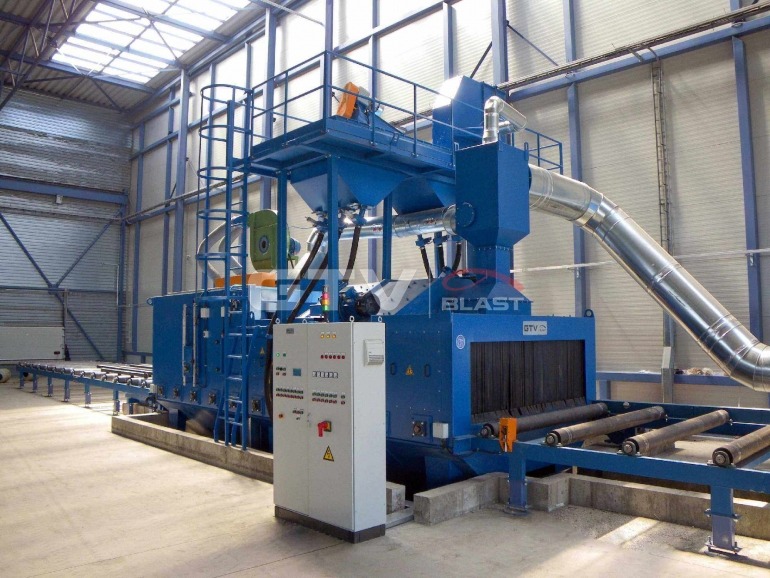 Niebieska maszyna do obróbki strumieniowo-ściernej w hali przemysłowej, z widocznymi rolotokami transportowymi i panelem sterowania z przyciskami oraz rurą odprowadzającą pył.