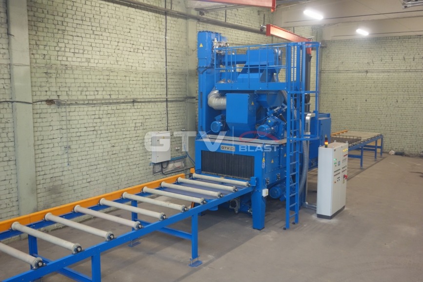 Niebieska maszyna do obróbki strumieniowo-ściernej z systemem transportu rolkowego w hali z ceglanymi ścianami, widoczna szafa sterownicza.