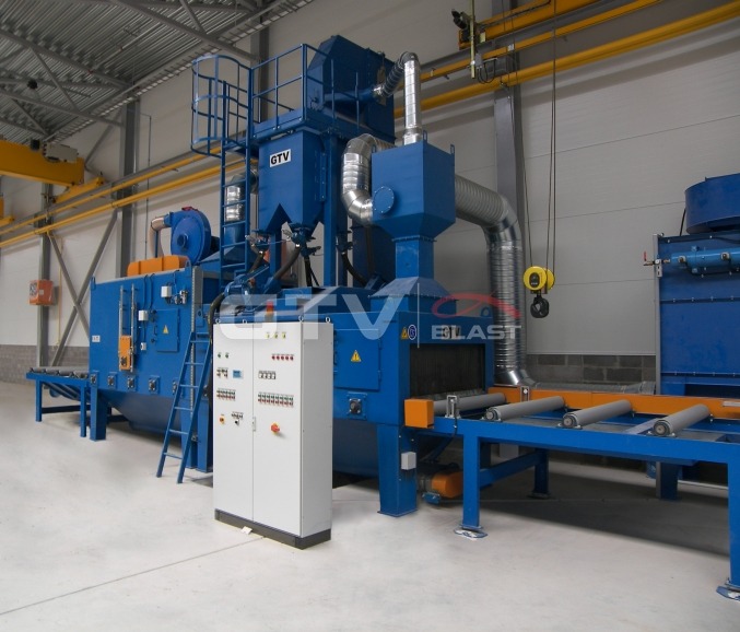 Kompleksowa, niebieska linia produkcyjna do obróbki strumieniowo-ściernej z widocznym panelem sterowania, transportem rolkowym i elementami wentylacyjnymi w hali przemysłowej.