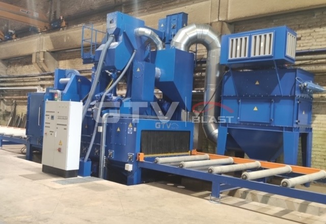 Kompleksowa, niebieska maszyna do obróbki strumieniowo-ściernej z panelami sterowania i systemem transportu rolkowego w hali przemysłowej.