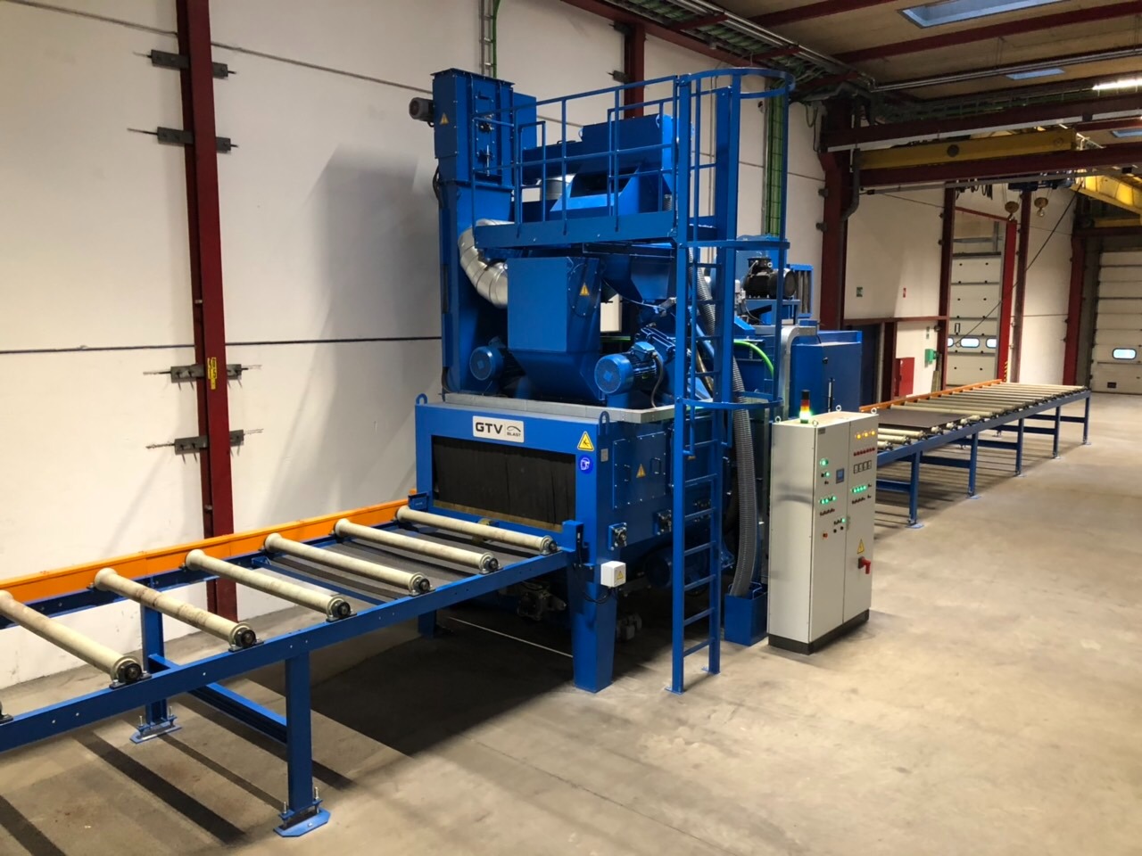 Niebieska oczyszczarka przelotowa z przenośnikiem rolkowym w hali produkcyjnej, widoczna drabina i panel sterowania, elementy konstrukcyjne pomalowane na czerwono