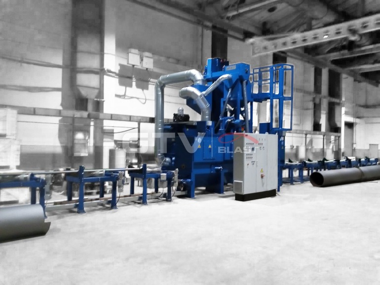 Niebieska oczyszczarka do rur w hali przemysłowej, z widocznym panelem sterowania i rurami na podajniku rolkowym.