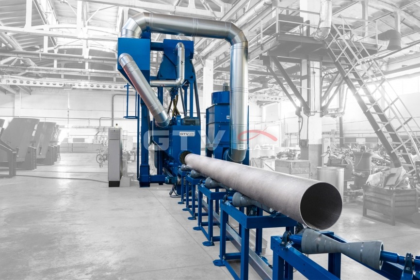 Wnętrze hali produkcyjnej z niebieską maszyną do obróbki strumieniowo-ściernej rur. Rura umieszczona na niebieskich rolkach transportowych, z widocznymi przewodami i elementami konstrukcyjnymi...