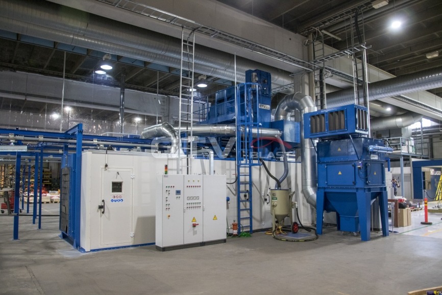 Oczyszczarka tunelowa z przenośnikiem podwieszonym w hali przemysłowej, widoczne elementy sterownicze, rury wentylacyjne i drabina dostępu do górnej części maszyny. Podłoga betonowa, oświetlenie...