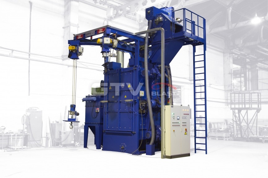 Niebieska śrutownica zawieszkowa z łańcuchem i hakiem w hali przemysłowej, widoczna drabina i panel sterowania.