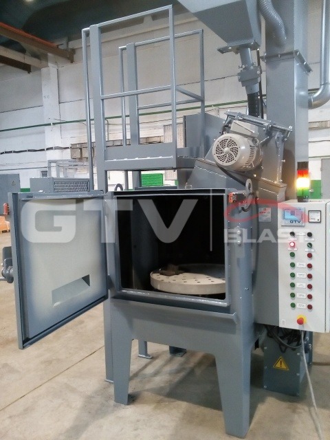 Szara śrutownica wirnikowa stołowa z otwartymi drzwiami, widoczny obrotowy stół wewnątrz komory, panel sterowania z przyciskami i lampkami kontrolnymi po prawej stronie, logo firmy na obudowie.