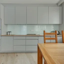 Jasnoszara kuchnia na wymiar z drewnianym blatem, białymi sprzętami AGD marki Beko i drewnianym stołem z krzesłami, oświetlona naturalnym światłem z okna z widokiem na budynek.