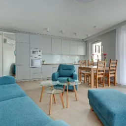 Jasne, przestronne wnętrze mieszkania z turkusową sofą, fotelem i pufem. W tle widoczna kuchnia z białymi szafkami i drewnianym blatem oraz stół z krzesłami z jasnego drewna.