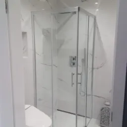 Biała łazienka z kabiną prysznicową narożną, wykończona marmurowymi płytkami, z widoczną toaletą myjącą i dekoracyjnym lampionem.