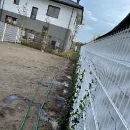 Nowo posadzony trawnik z widocznym systemem nawadniania i ogrodzeniem panelowym, na tle nowoczesnego domu.