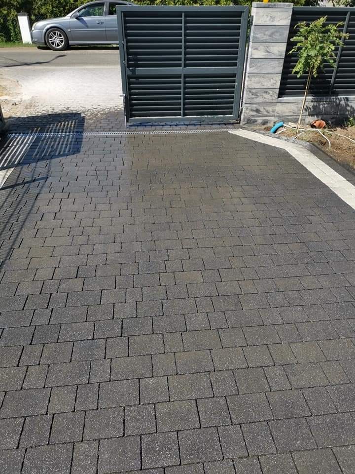 Ułożona, grafitowa kostka brukowa na podjeździe, z widoczną bramą przesuwną w tle i fragmentem szarego samochodu osobowego po lewej stronie.