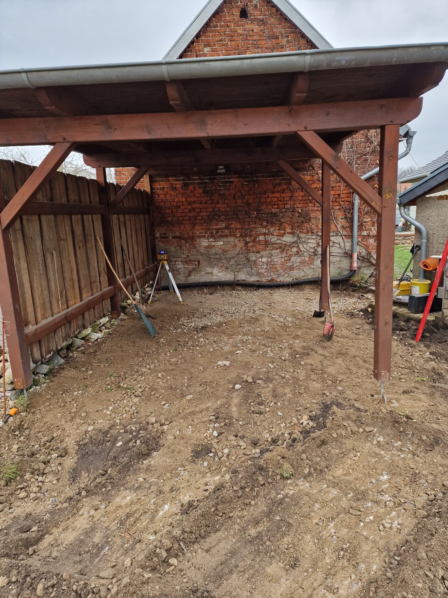 Przygotowanie terenu pod konstrukcję drewnianą: wyrównana ziemia z kamieniami, widoczne narzędzia (łopata, poziomica laserowa) oraz drewniany szkielet z zadaszeniem na tle ceglanego budynku.