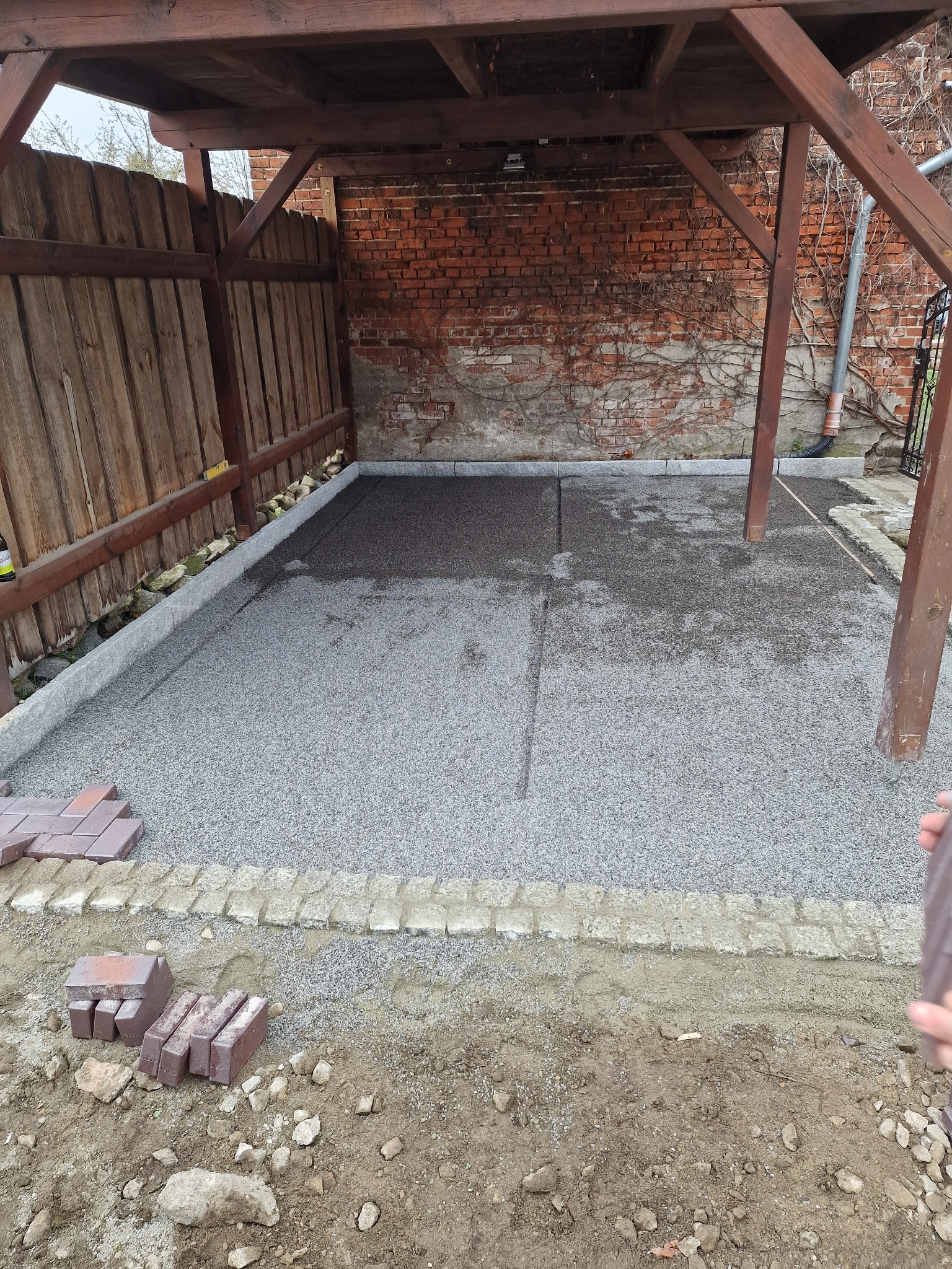 Przygotowanie podłoża pod zadaszeniem: warstwa podsypki z grysu, obrzeża z kostki granitowej i stos cegieł brukowych czekających na ułożenie.