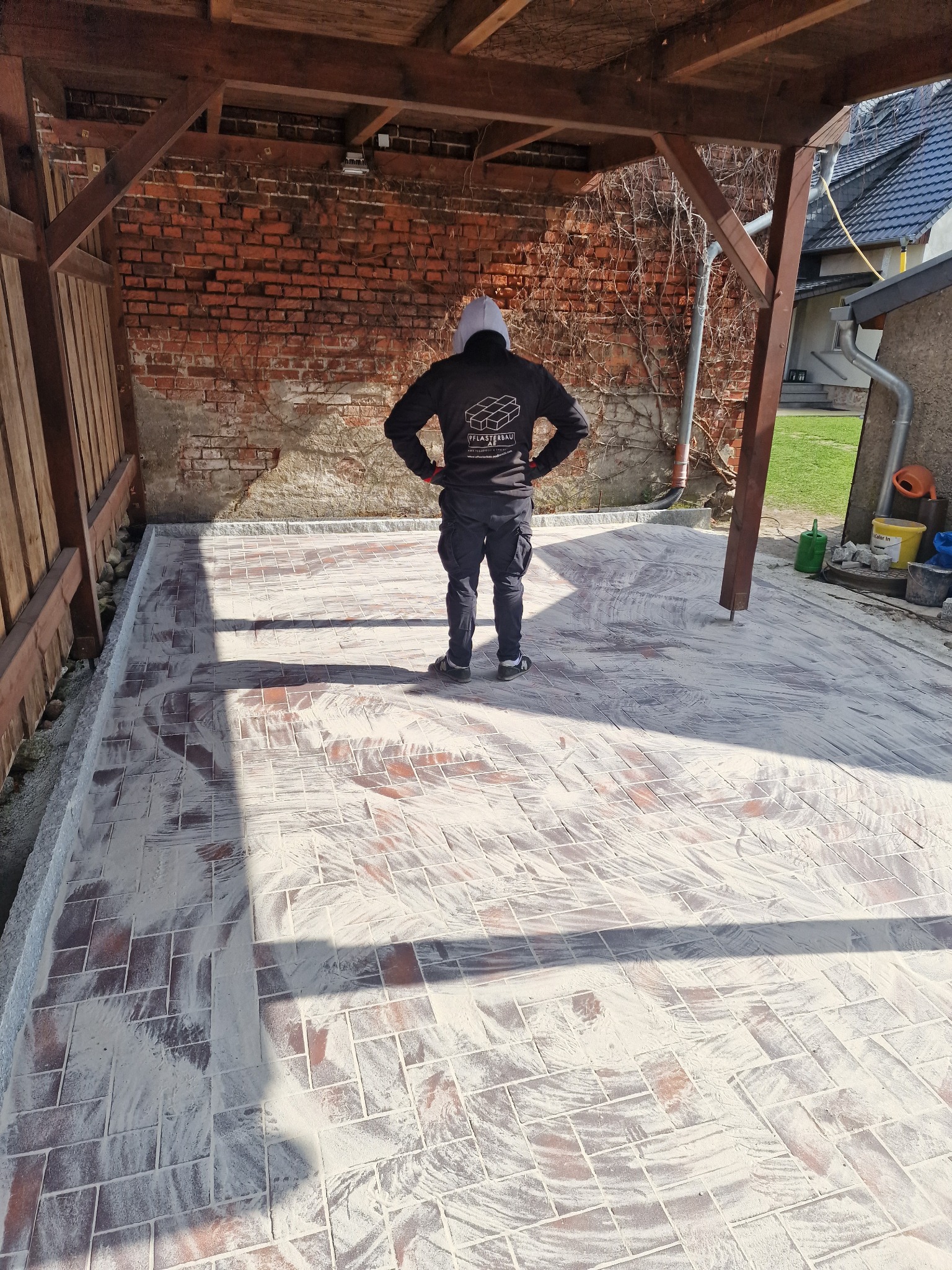 Układanie kostki brukowej pod drewnianą pergolą, widok zza pleców osoby nadzorującej prace, kostka zasypana piaskiem kwarcowym, mur z czerwonej cegły w tle.