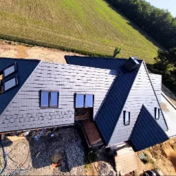 Nowoczesny dom z ciemnoszarym dachem łupkowym, widok z góry na tle zielonego wzgórza. Dach z oknami połaciowymi i kominem. Częściowo wykończone otoczenie domu.