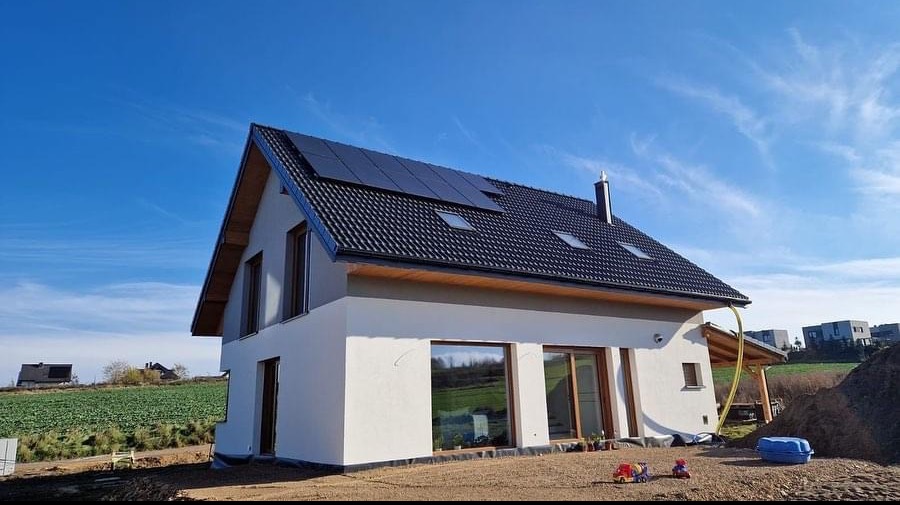 Nowoczesny dom jednorodzinny z czarną dachówką i panelami słonecznymi, widoczny z zewnątrz w słoneczny dzień, z elementami wykończeniowymi i zabawkami dziecięcymi na pierwszym planie.