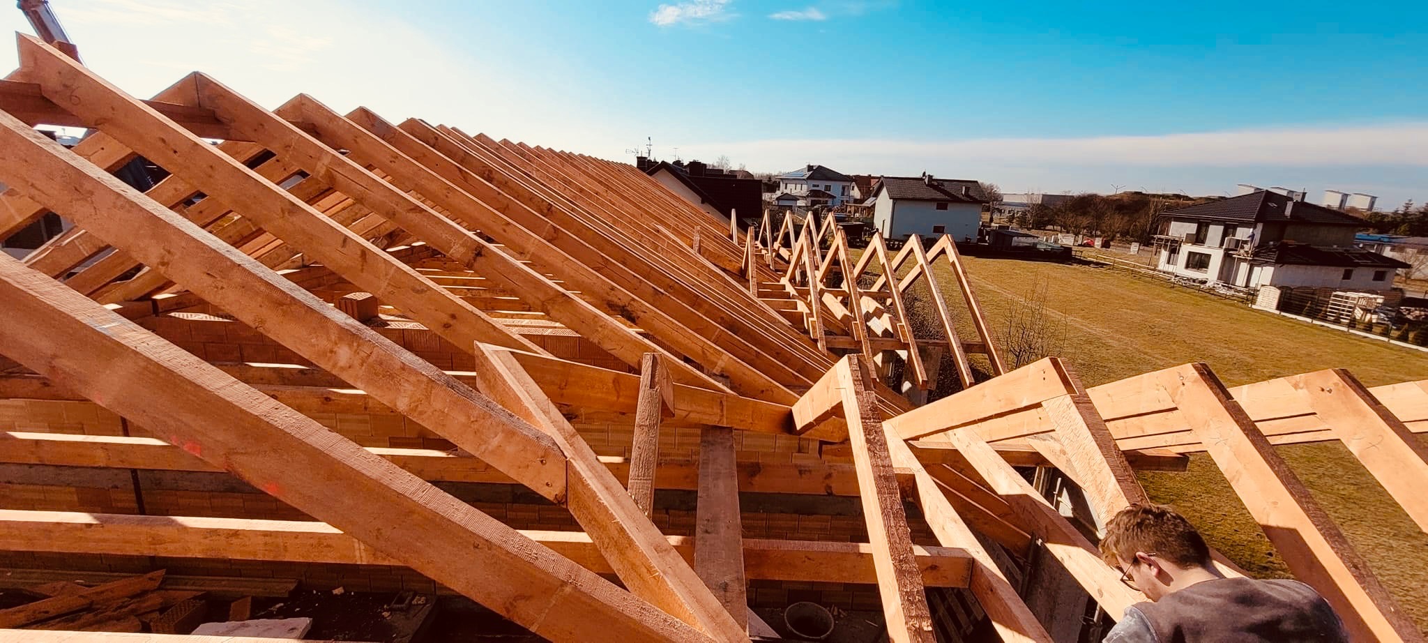 Wysoka perspektywa na drewnianą konstrukcję dachu w trakcie budowy, z widocznym pracownikiem w okularach i zabudową mieszkalną w tle pod błękitnym niebem.