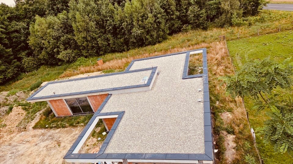 Nowoczesny dom z płaskim dachem pokrytym jasnym żwirem, widok z góry, obrzeża dachu wykończone ciemną blachą, w tle zieleń drzew i trawy.