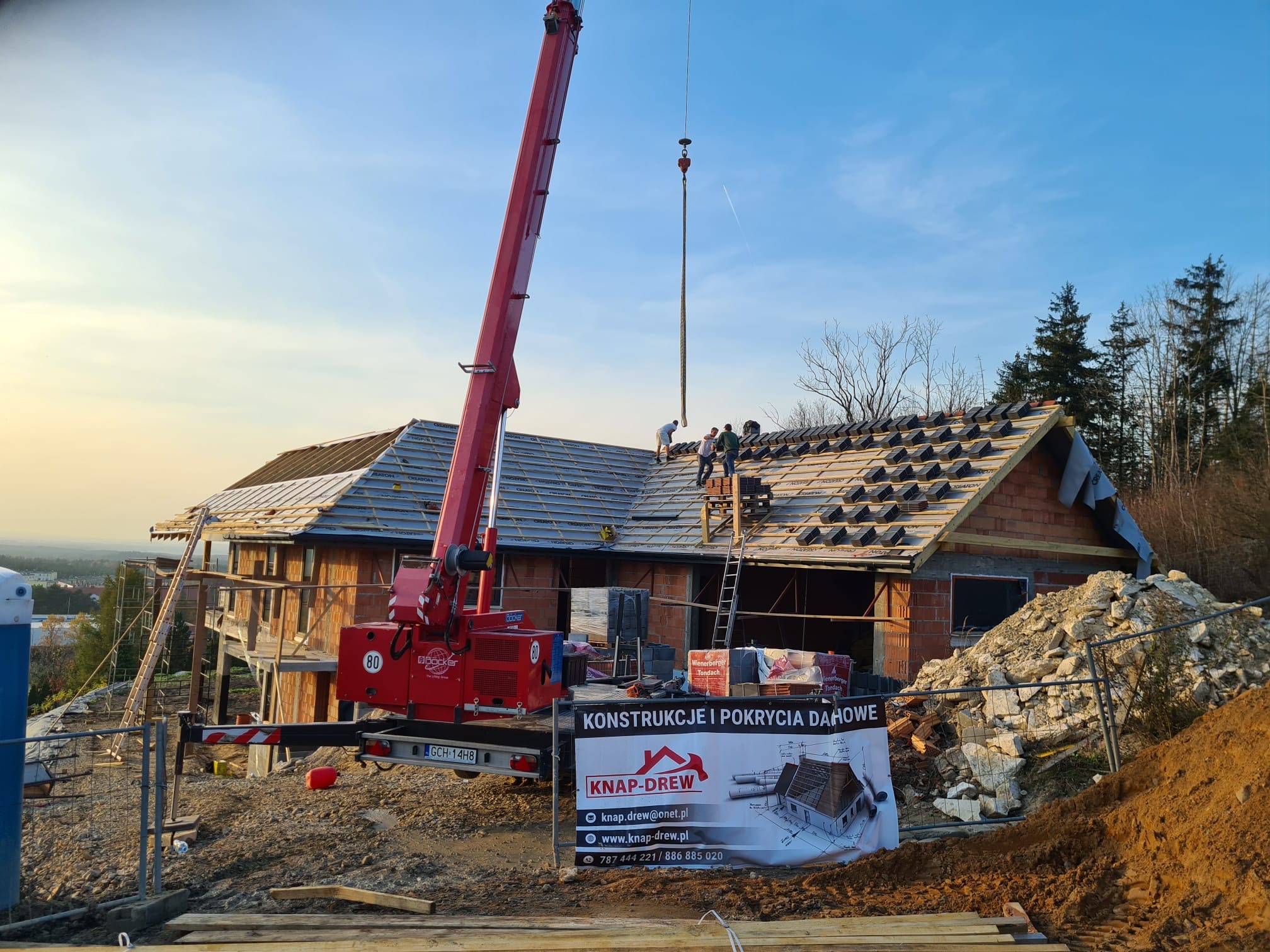 Montaż dachówki na nowym domu jednorodzinnym z użyciem dźwigu, widoczna konstrukcja więźby i ekipa dekarzy pracująca na dachu, w tle las i błękitne niebo.