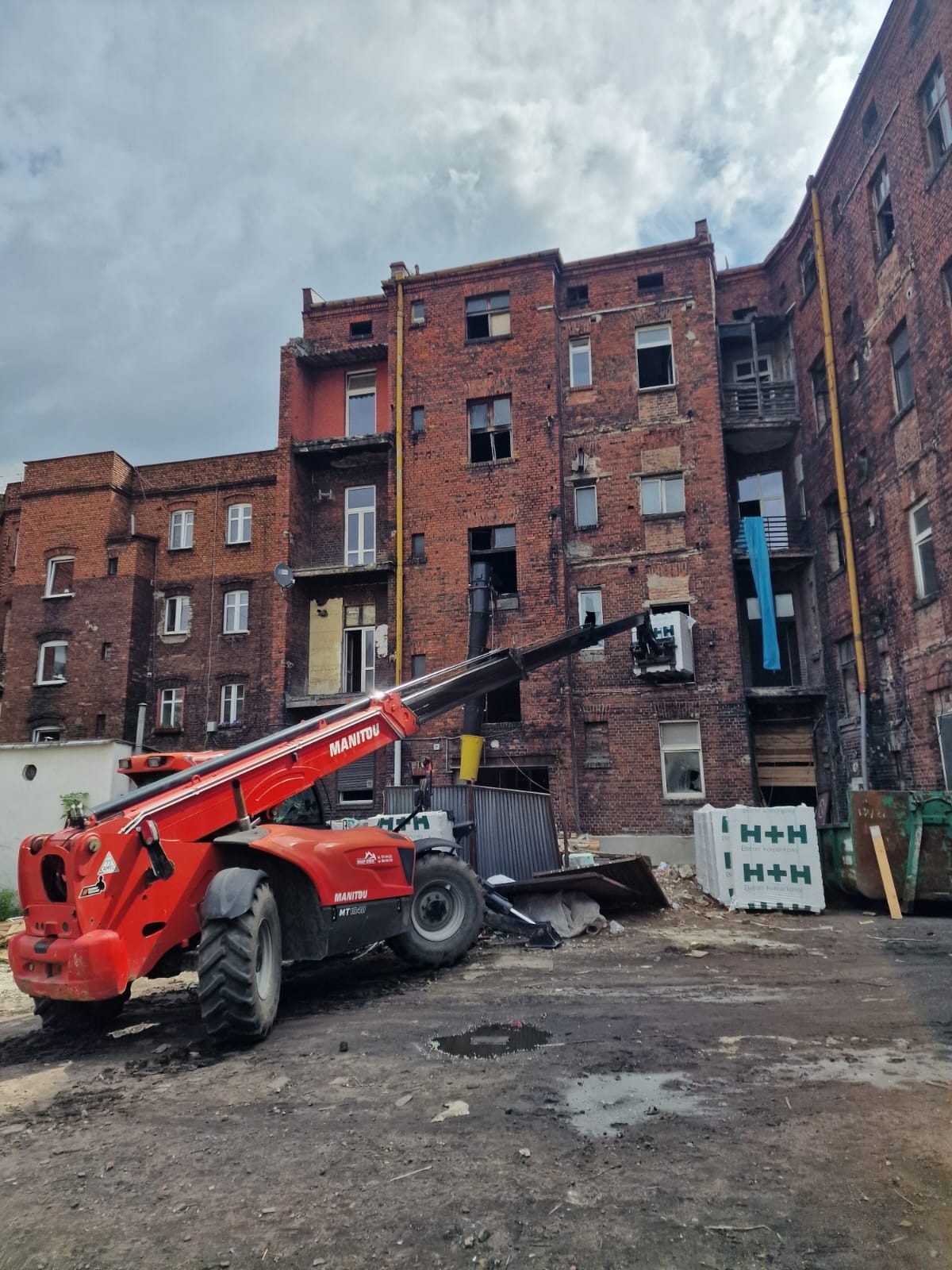 Czerwony podnośnik Manitou na placu budowy przy ceglanym budynku z uszkodzonymi oknami. Materiały budowlane i kontenery na gruz w tle.
