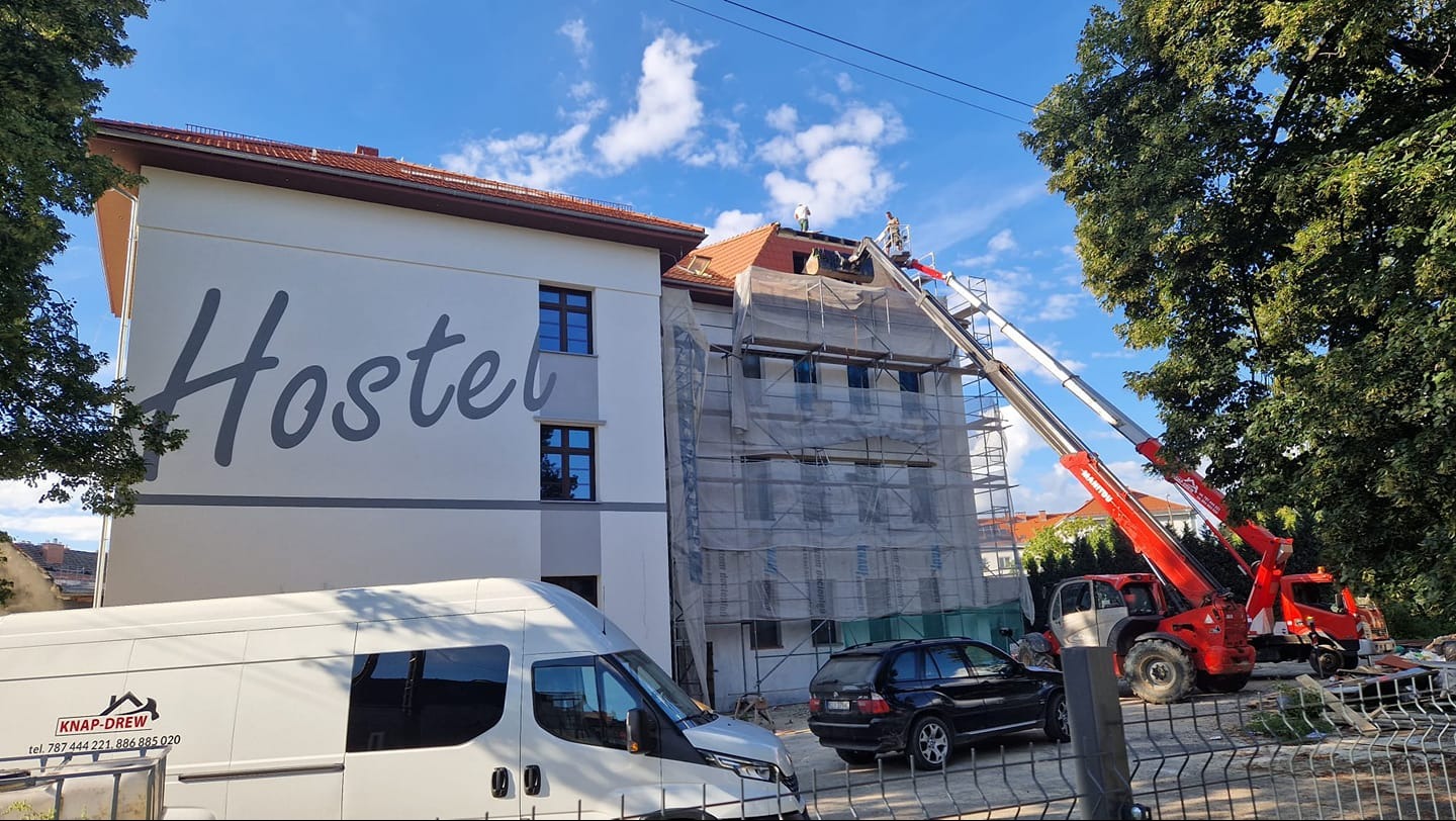 Remont dachu budynku z napisem 'Hostel', widoczny z bliska podnośnik koszowy i rusztowania. Częściowo zasłonięty samochód dostawczy i osobowy.