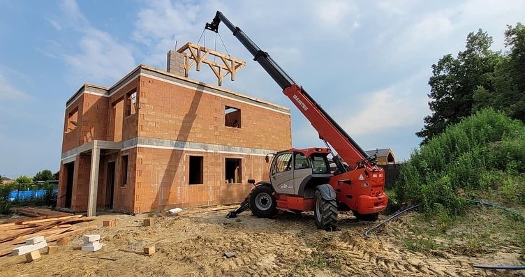 Czerwony dźwig MANITOU podnosi drewnianą konstrukcję więźby dachowej na budynek z czerwonej cegły w trakcie budowy, widok na zielone wzgórze w tle.