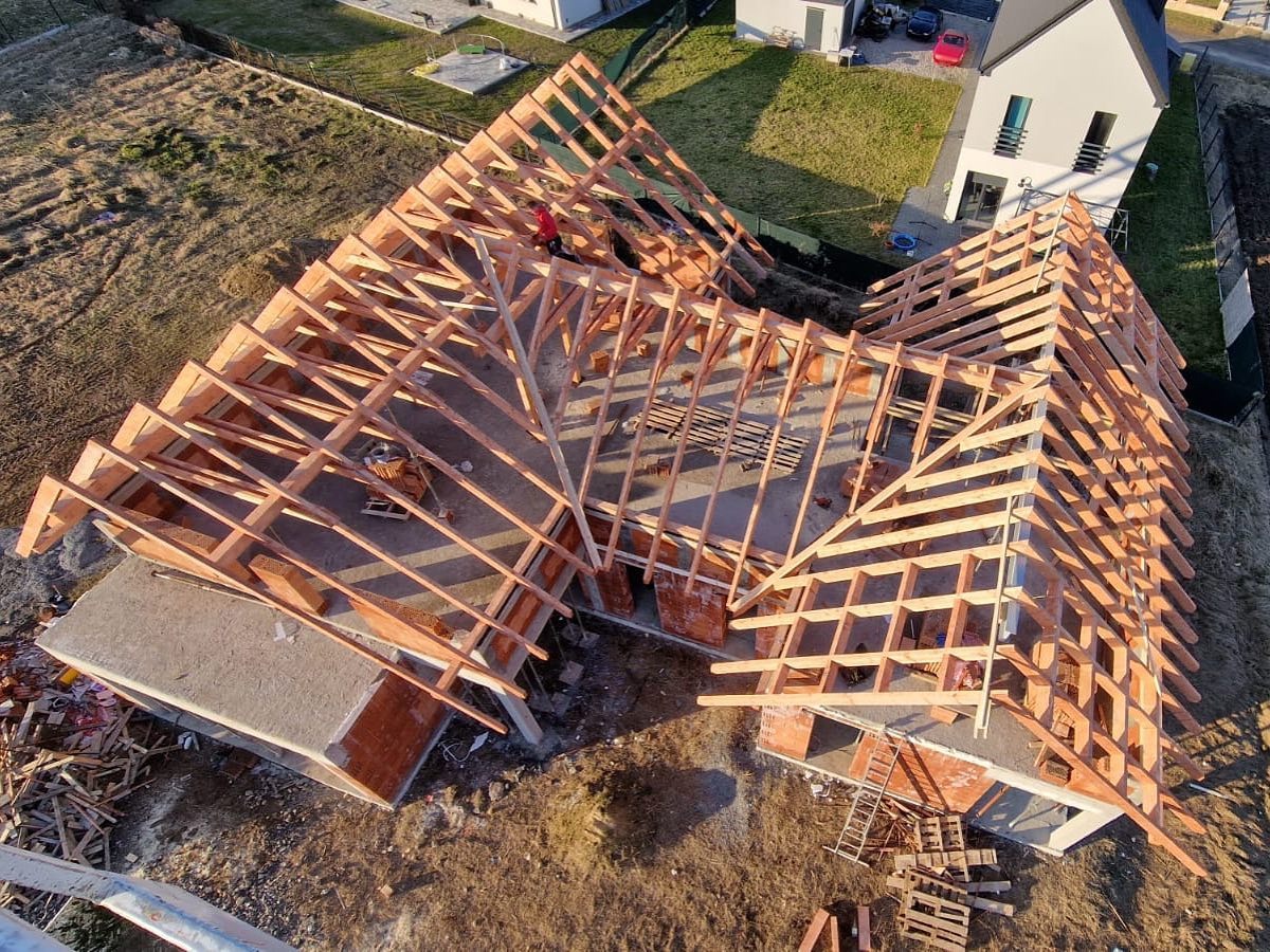 Wysoka perspektywa na budowę dachu domu jednorodzinnego z widoczną drewnianą konstrukcją więźby i pracującym dekarzem w czerwonej kurtce. Słoneczny dzień.