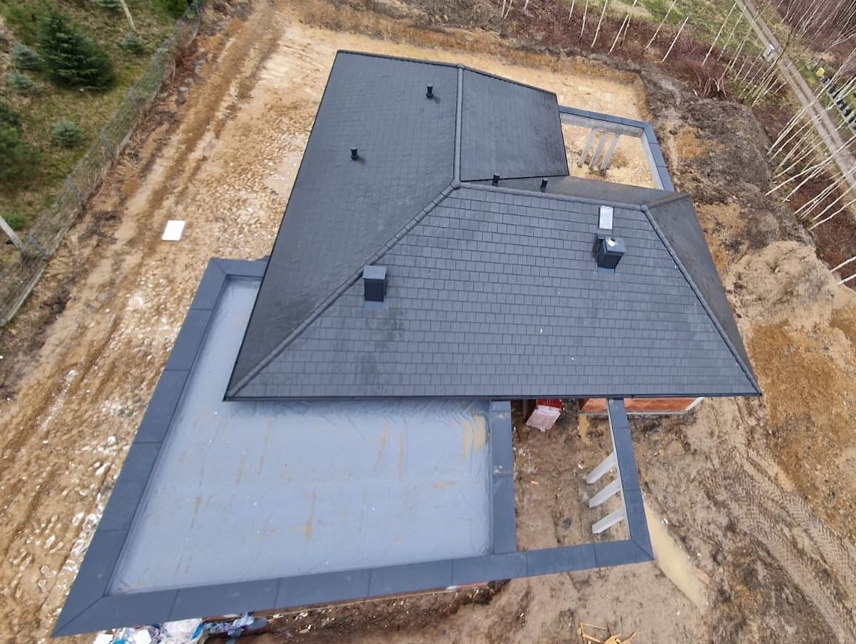 Widok z góry na ciemnoszary dach z dachówki bitumicznej na domu w trakcie budowy, z widocznymi elementami konstrukcyjnymi i otoczeniem placu budowy.