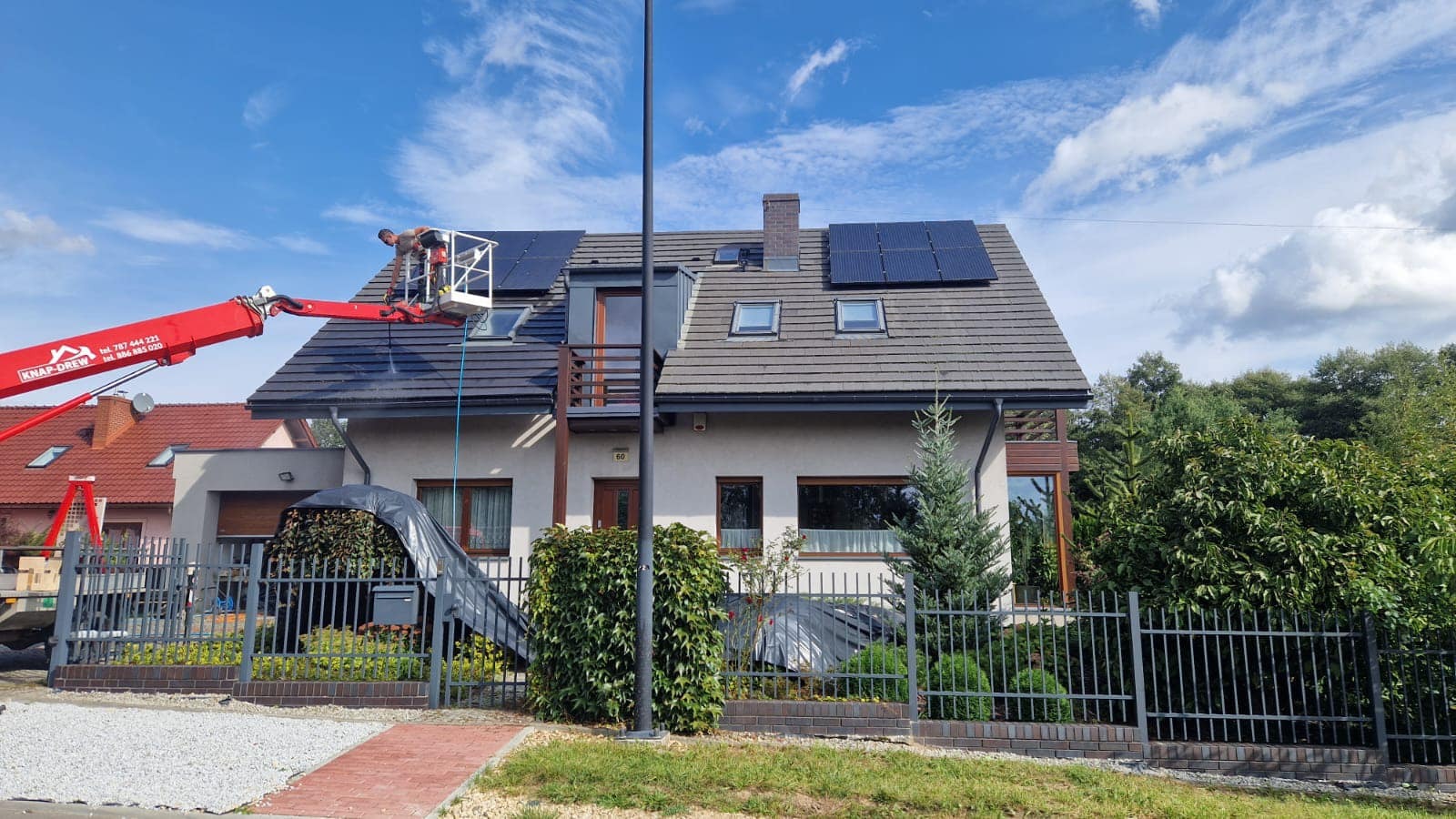 Czyszczenie dachu z paneli solarnych przez pracownika na podnośniku, z użyciem wody pod ciśnieniem. Widoczny dom z szarą dachówką i panelami.
