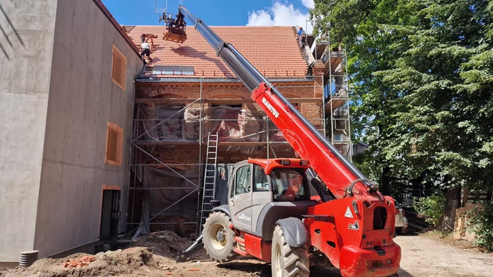 Czerwony podnośnik Manitou unosi materiały na dach remontowanego budynku z cegły, gdzie pracują dekarze. Na pierwszym planie widoczny fragment otynkowanej ściany.